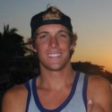 Kyle Liddell, smiling in a tank top and backwards cap, stands outdoors at sunset with palm trees and buildings silhouetted against the vibrant sky.