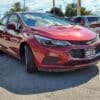 A 2018 Chevrolet Cruze LT Turbo sedan in red is parked in a lot under a partly cloudy sky, with several other vehicles visible in the background, and it has a dealership plate on the front.