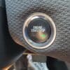 Close-up of a textured dashboard from a 2018 Chevrolet Cruze LT Turbo, featuring a circular engine start/stop button with a chrome rim. A small green light is visible below the button, along with part of a shoe at the bottom.
