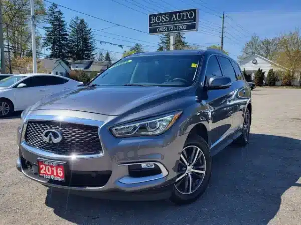 A 2016 gray Infiniti QX60 AWD is parked in a lot beneath a Boss Auto sign, sporting a red dealership plate. Framed by other cars, trees, and a partly cloudy sky, this SUV captures attention.