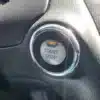 A close-up of the 2016 Infiniti QX60 AWD engine start-stop button shows a circular design with a metallic ring and black surroundings, featuring START STOP ENGINE text and an orange indicator above it, highlighting its sleek design.