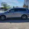 A 2016 Infiniti QX60 AWD glistens in the sun, poised for adventure. Parked among vibrant trees and charming houses, the silver SUV showcases its readiness against a picturesque backdrop.