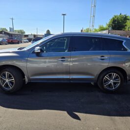 A 2016 Infiniti QX60 AWD is parked in a sunny lot alongside other cars, with a clear blue sky above and trees and buildings visible in the distance.