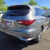 A 2016 Infiniti QX60 AWD, silver, is parked on asphalt in a lot surrounded by other cars and greenery, seen from the rear left under a clear blue sky. A dealer plate reading “Loaded” is visible.