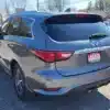 A 2016 Infiniti QX60 AWD in gray is parked with its rear end facing the camera, showcasing taillights and rear window. Its license plate frame displays Loaded Boss Auto. Trees and houses appear in the background beneath a clear sky.