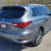 A silver 2016 Infiniti QX60 AWD SUV with a LOADED dealership plate is parked in a sunny lot. Its rear and side are visible, amidst other cars and bare trees.