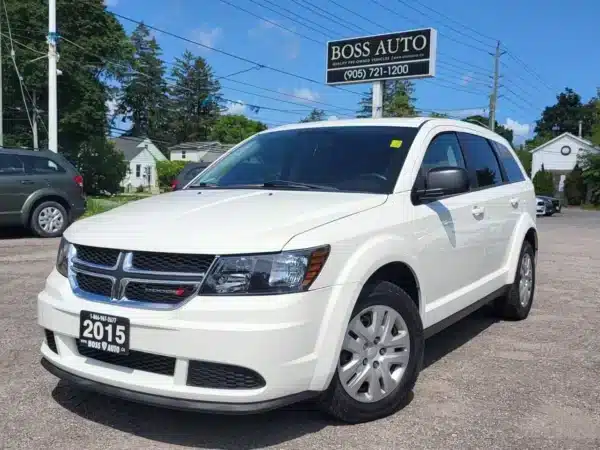 A 2015 Dodge Journey SE in white gleams under the sun at a dealership, with a BOSS AUTO sign displaying contact details nearby. The license plate highlights its vintage, framed by distant trees and buildings.