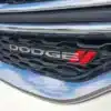 Close-up of the 2015 Dodge Journey SE grille, featuring Dodge in silver and a red logo on a honeycomb background. Chrome and black detailing frame it, with reflections of blue sky and clouds shimmering across the surface.