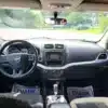 From the drivers seat of the 2015 Dodge Journey SE, you see the dashboard with a central screen, air vents, and various controls. Two Thank You floor mats are present. The green landscape is visible through the windshield.