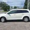 A 2015 Dodge Journey SE, in white, is parked sideways in a lot on a sunny day, highlighting its length. Houses, trees, and power lines form the scenic backdrop.