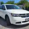 A white 2015 Dodge Journey SE is parked on a gravel lot, featuring a front plate reading 2015. Trees and part of a building with an Auto Sale sign are visible in the background.