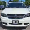 A 2015 Dodge Journey SE, white in color, is parked among other vehicles in a car lot. Its front view showcases the distinct Dodge grille and headlights against a backdrop of trees. A dealership sign on the front bumper accentuates its sleek design.