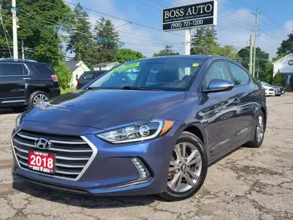 A metallic blue 2018 Hyundai Elantra Limited with a Boss Auto license plate is parked on a gravel lot. A dealership sign, trees, and other vehicles are visible in the background.