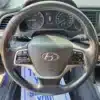 Close-up of a 2018 Hyundai Elantra Limited steering wheel featuring audio and phone controls. The dashboard includes a speedometer and fuel gauges, while the multimedia display reads 99.9. A partially visible sign in the background enhances the sleek interior of this model.