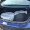 The open trunk of a 2018 Hyundai Elantra Limited reveals two large white plastic-wrapped items, partially covered by a black car floor mat. Parked on a paved surface, the blue vehicle has a backdrop of grass.