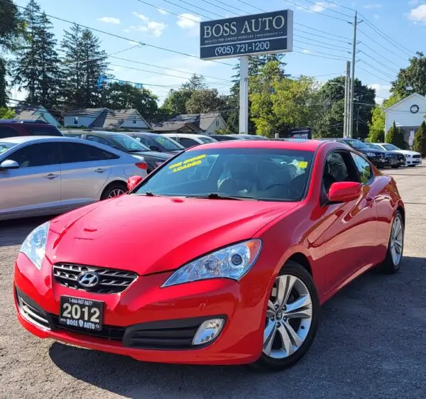 A red 2012 Hyundai Genesis 2.0T Premium is parked at a used car dealership with a yellow SALE sticker on the windshield. Several other cars and a Boss Auto sign with a phone number can be seen in the background.