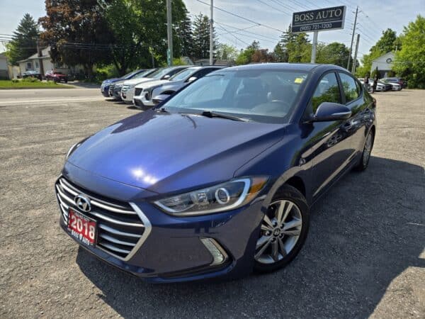 A 2018 Hyundai Elantra Limited is parked at a car dealership lot under sunny skies, with other vehicles and a BOSS AUTO sign visible in the background.