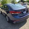 A 2018 Hyundai Elantra Limited in dark blue is parked in a sunlit lot, seen from the rear left. A GAS SAVER dealership plate is on the back, with trees and other cars visible in the background.