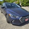 A 2018 Hyundai Elantra Limited sedan in blue is parked on a gravel lot with its front wheels turned, surrounded by other vehicles and trees in the background.