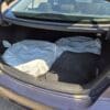 The open trunk of a 2018 Hyundai Elantra Limited (dark blue) contains two large tires in white plastic bags and a black floor mat labeled Elantra. The trunk is mostly empty, with sunlight illuminating the scene.