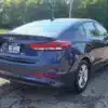 A 2018 Hyundai Elantra Limited is parked in a sunlit area with trees behind. The rear view highlights its sleek tail lights and design, with the license plate GAS SAVER.