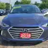 A blue 2018 Hyundai Elantra Limited sedan is parked in a lot with an EXECUTIVE sticker on the windshield, a yellow tag on the hood, and a red license plate featuring 2018 and a phone number. Other cars are visible under the clear sky in the background.
