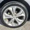 The wheel of a 2014 Hyundai Elantra GT is captured on a sunny day, featuring a silver alloy, five-spoke rim, fitted with a Nexen tire. It is elegantly parked on the textured asphalt.