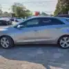 A 2014 Hyundai Elantra GT gleams with its silver finish on the asphalt among other vehicles, reflecting the clear sky. In the background, trees sway gently near charming houses, crafting an idyllic parking lot scene.