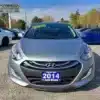 A 2014 Hyundai Elantra GT in silver is parked among other vehicles. The Boss Auto dealership name and a green windshield sticker are visible, with trees and a clear sky as the scenic backdrop.