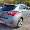 A silver 2014 Hyundai Elantra GT hatchback is parked on gravel, showcasing tinted rear windows and a sales ad placeholder plate. Trees and other vehicles in the background add depth to the scene.