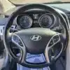 Close-up of the 2014 Hyundai Elantra GT steering wheel with control buttons. The dashboard shows speed and RPM dials alongside digital info, while a partially open door hints at the Elantra GTs sleek interior design.