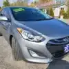 A silver 2014 Hyundai Elantra GT shines in front of Auto Sales, with a LOW MILES sticker on the windshield, as trees sway gently behind it.