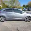 A sleek 2014 Hyundai Elantra GT hatchback is parked side-on in the lot, surrounded by several vehicles. Trees and a small building under a clear sky create a picturesque backdrop.