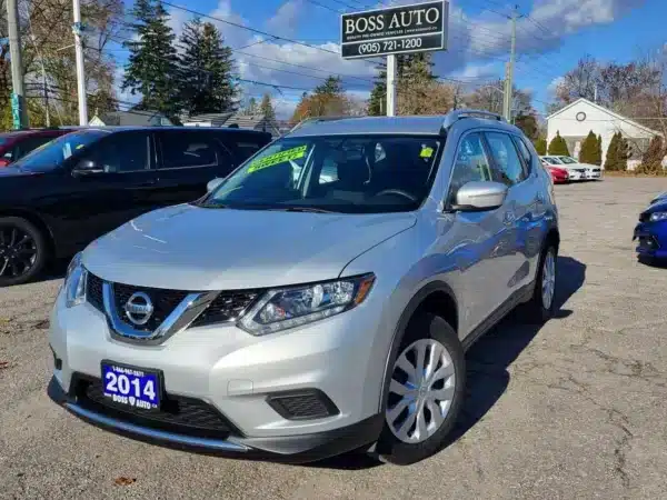 A 2014 Nissan Rogue 2.5 SV SUV in silver is parked at a car dealership with a Boss Auto sign and phone number visible. A bright green label on the windshield showcases a special feature, while other vehicles wait nearby for eager buyers.