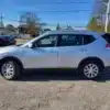 A silver 2014 Nissan Rogue 2.5 SV is parked on an asphalt lot with other vehicles in the background, under a clear blue sky, with trees and houses visible behind the lot.