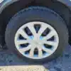 Close-up of a 2014 Nissan Rogue 2.5 SV tire on pavement. The black tread shows visible text patterns, paired with a silver alloy wheel featuring a unique spoke design. Part of the cars sleek body is seen in the top left.