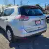 A silver 2014 Nissan Rogue 2.5 SV is parked in a sunny lot with a Certified sticker on its license plate, a black car partially visible nearby, and serene trees and houses in the background.