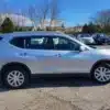 A silver 2014 Nissan Rogue 2.5 SV is parked on a paved lot with several other cars, set against a backdrop of sparsely leaved trees and houses under a partly cloudy sky.