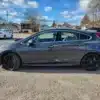 On a sunny day, a dark gray 2017 Chevrolet Cruze Premier Hatchback with black rims and red brake calipers is parked in a residential area. Houses, trees, and other parked cars are in the background under a blue sky with clouds.