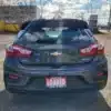 The image shows the rear of a 2017 Chevrolet Cruze Premier Hatchback in dark gray, parked in a lot. Featuring distinctive taillights and a central logo, its Loaded license plate frame is from Boss Auto. Trees and buildings serve as the backdrop.