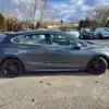 A gray 2017 Chevrolet Cruze Premier Hatchback, compact and with sleek black wheels, is parked in an outdoor lot. Its viewed from the side with other cars and leafless trees in the partly cloudy background.