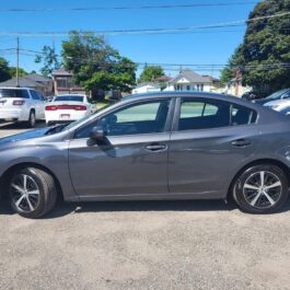 A 2019 Subaru Impreza 2.0i AWD sedan in gray is parked in a lot on a sunny day, surrounded by other cars, with houses and trees in the background.