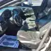 The 2015 Chrysler 200 S AWDs black interior features leather seats, a steering wheel, and a sleek dashboard. The drivers door is open, displaying an AWD paper mat with footprints, while sunlight beautifully highlights the intricate seat designs.
