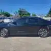 A 2015 Chrysler 200 S AWD is parked in a lot on a clear day. The black sedan, famous for its sleek design and AWD, stands out amid vehicles, houses, and trees beneath the bright blue sky.