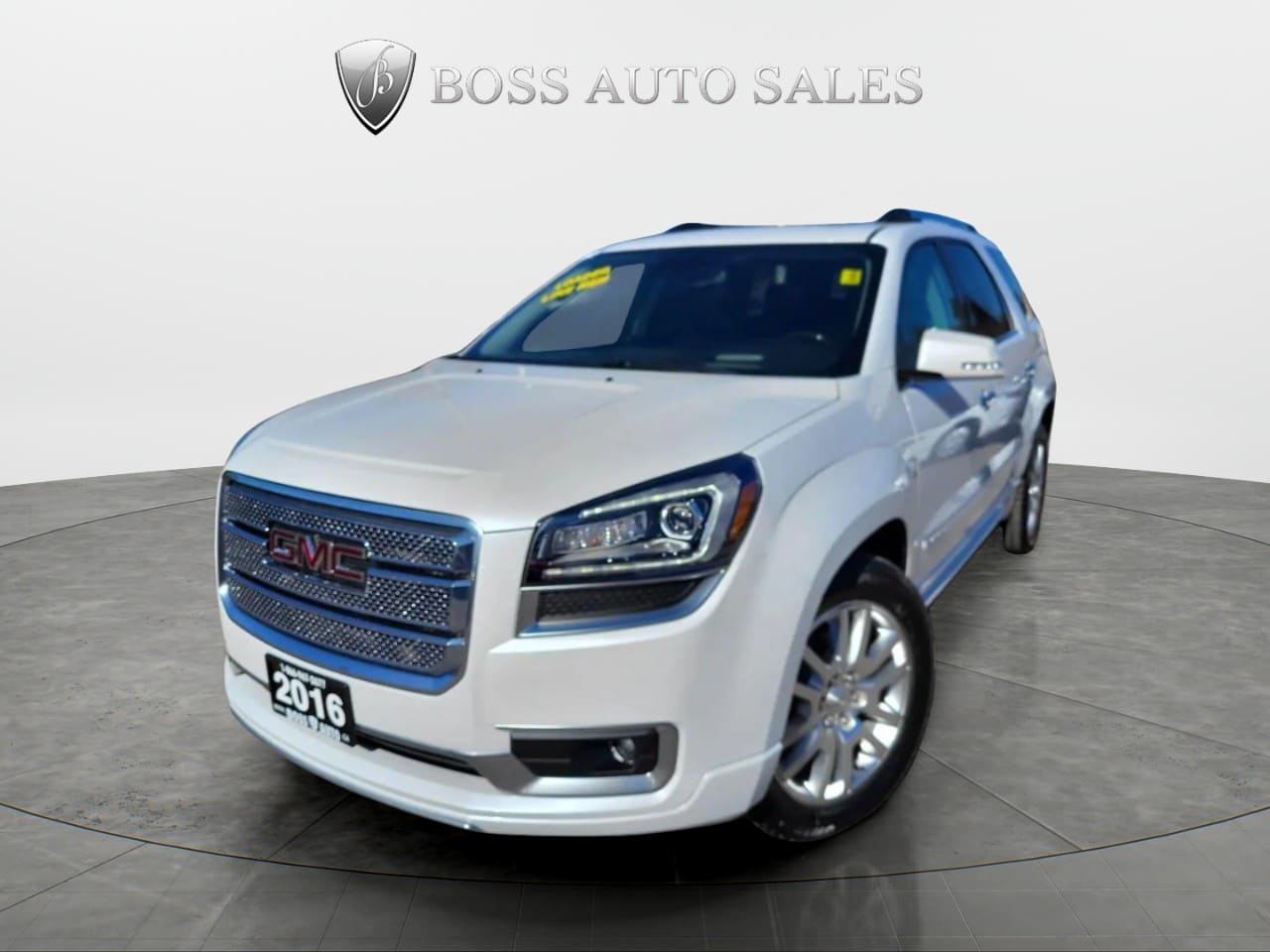 A 2016 GMC Acadia Denali AWD in white is showcased at an angle in a showroom, highlighting its front grille, headlights, and side profile with the BOSS AUTO SALES logo displayed on the wall behind it.