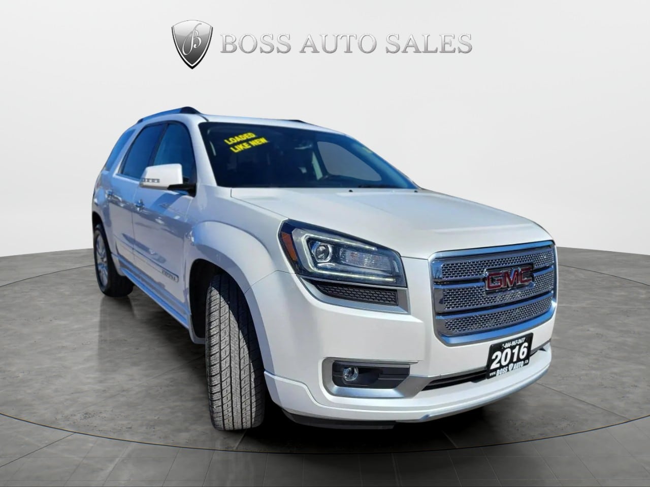 A white 2016 GMC Acadia Denali AWD SUV is displayed in a showroom beneath the Boss Auto Sales logo. A yellow "Loaded, Low Kms" sign is on the windshield, and the vehicle is angled to show its front and driver’s side.