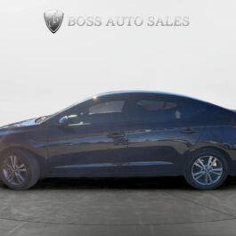 Side view of a black 2017 Hyundai Elantra SE sedan parked indoors on a gray tiled floor, with the Boss Auto Sales logo and text displayed on the white showroom wall. Side view of a black 2017 Hyundai Elantra SE sedan parked indoors on a gray tiled floor, with the Boss Auto Sales logo and text displayed on the white showroom wall.