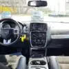 The 2017 Dodge Grand Caravan SXTs interior boasts a sleek steering wheel, advanced dashboard with touchscreen console, and intuitive controls. The front seats offer comfort, while outside the windshield several parked cars can be seen in the background.