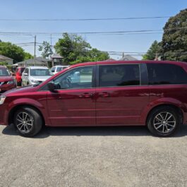 A red 2017 Dodge Grand Caravan SXT is parked in a lot on a sunny day, with other cars, trees, and houses visible in the background.