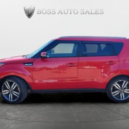 A 2015 Kia Soul SX Luxury, a red compact SUV with black trim and alloy wheels, is parked indoors against a white background. 